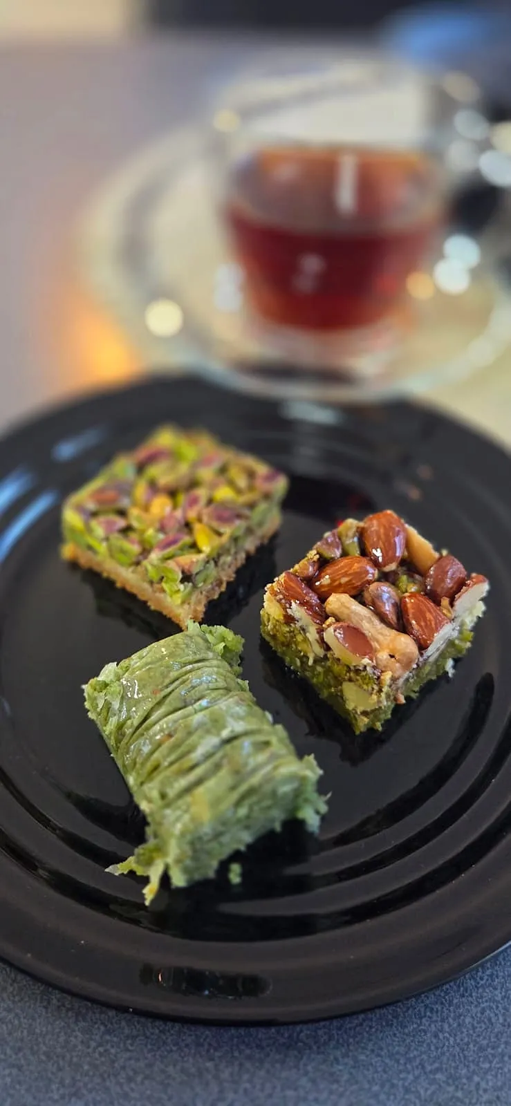 Drei Stück Pistazien-Baklava auf einem schwarzen Teller, im Hintergrund ein Glas Tee